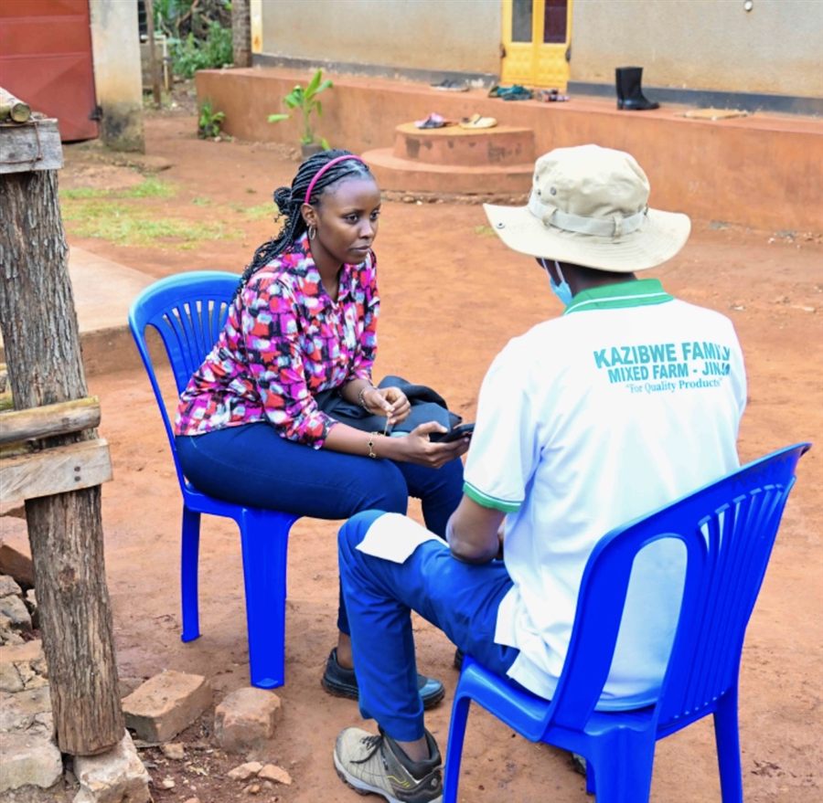 Kellen Owente seated outdoors, interviewing a Kazibwe Family Mixed Farm representative in Mayuge for a Parish Development Model field visit.